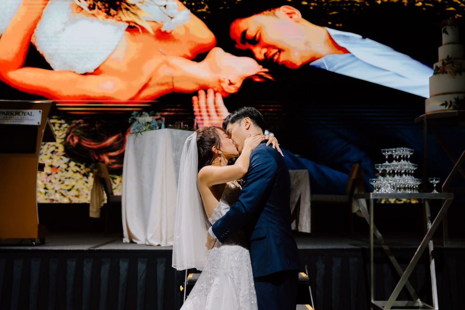 Bride and groom share first kiss at wedding reception with romantic photo display backdrop.