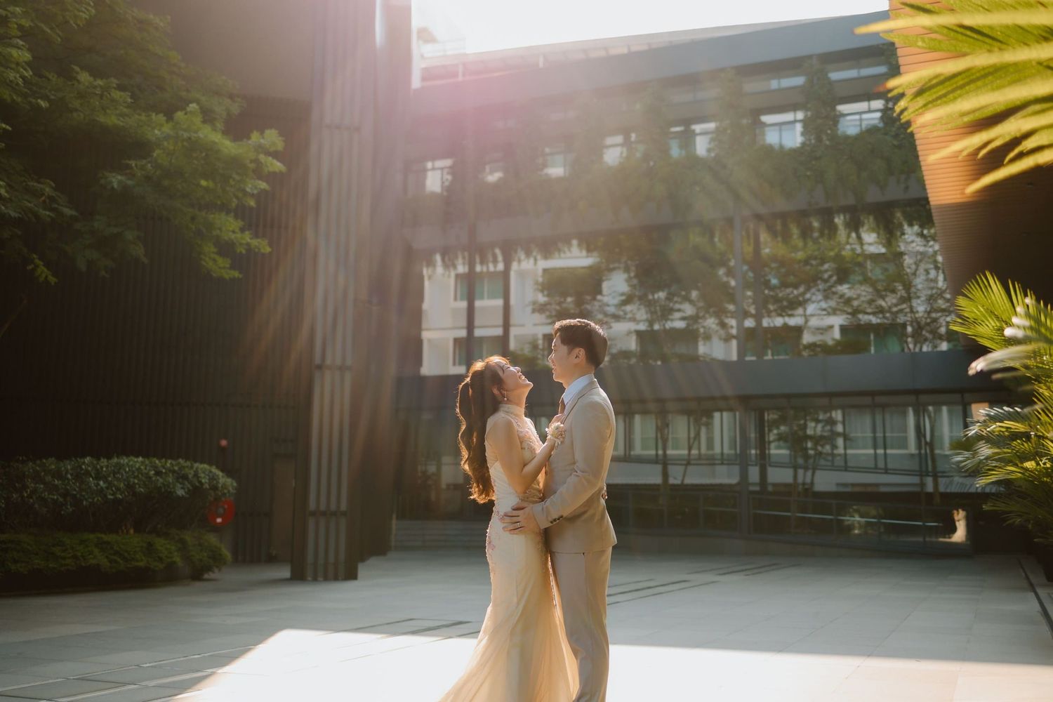 Couple embraces in golden sunset light outside modern building, bride in elegant gown and groom in suit.