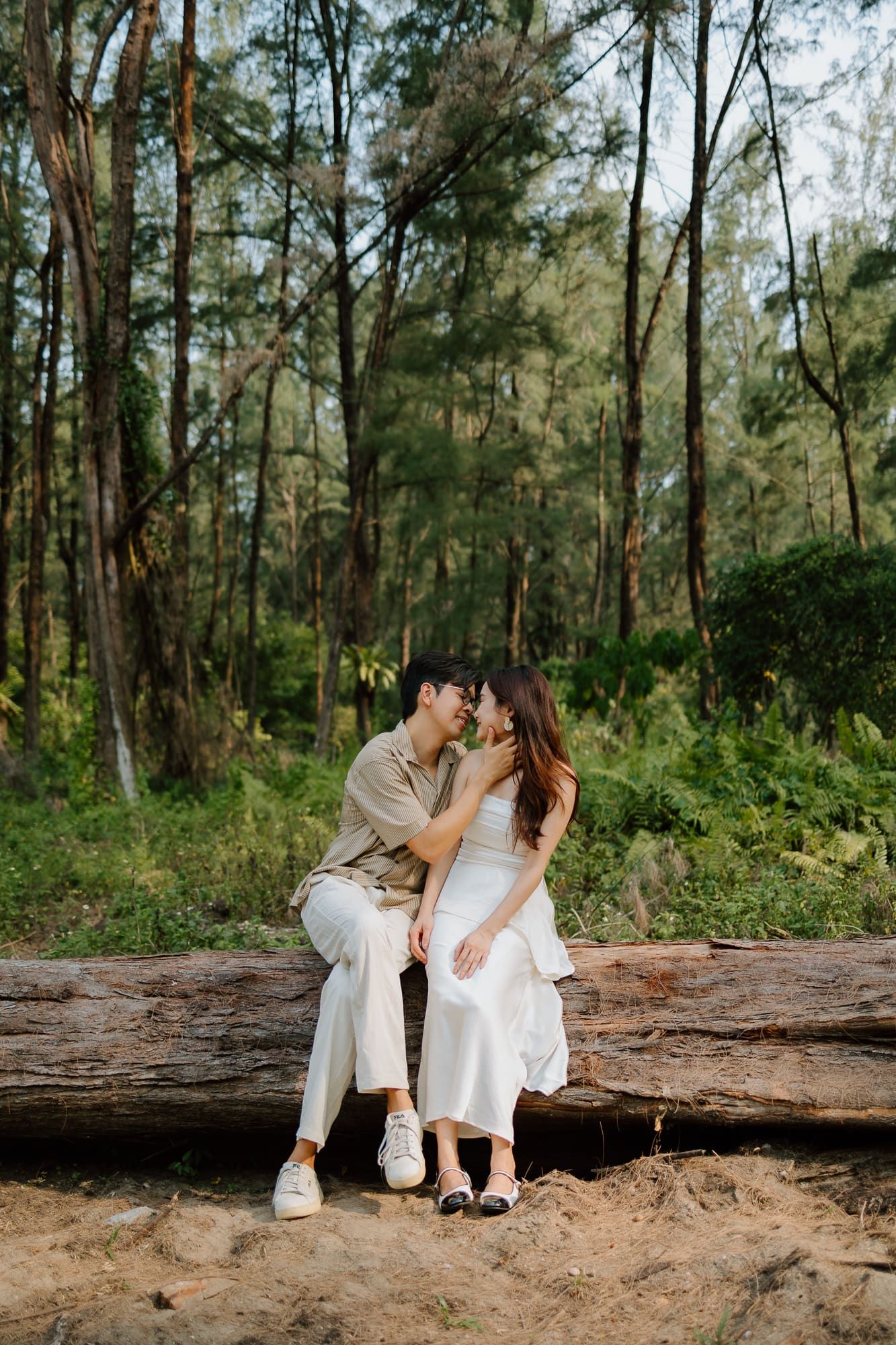 Couple sitting on a fallen log in a lush forest, sharing a romantic moment in neutral tones.
