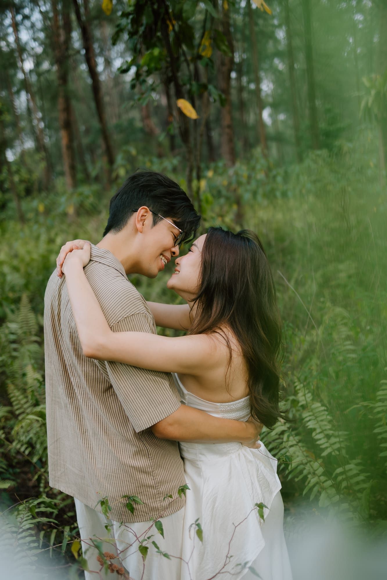 A couple embraces intimately in a lush green forest, sharing a romantic moment surrounded by ferns and trees.