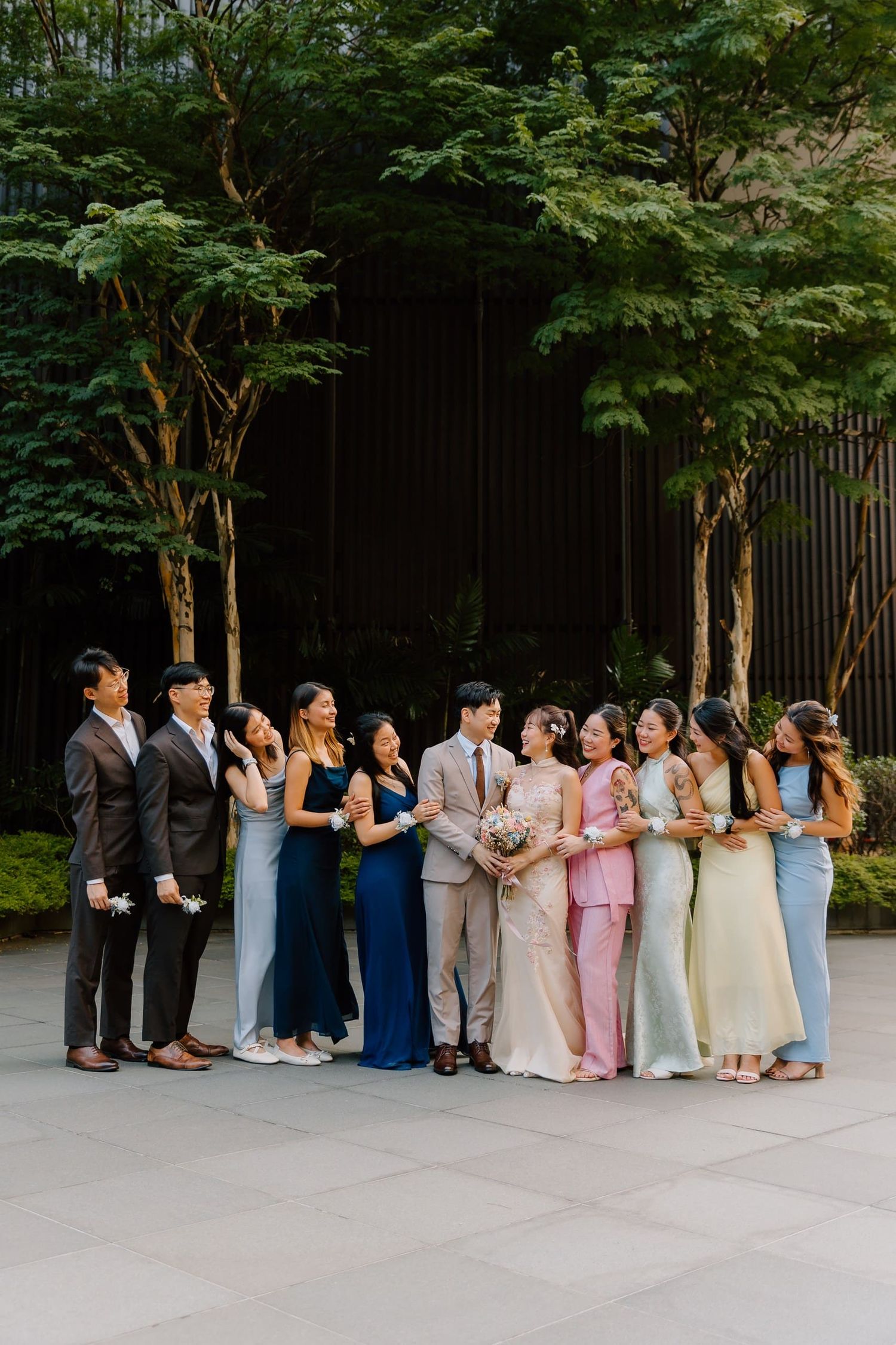 Wedding party in colorful gowns and suits pose together outdoors with lush green trees in background.