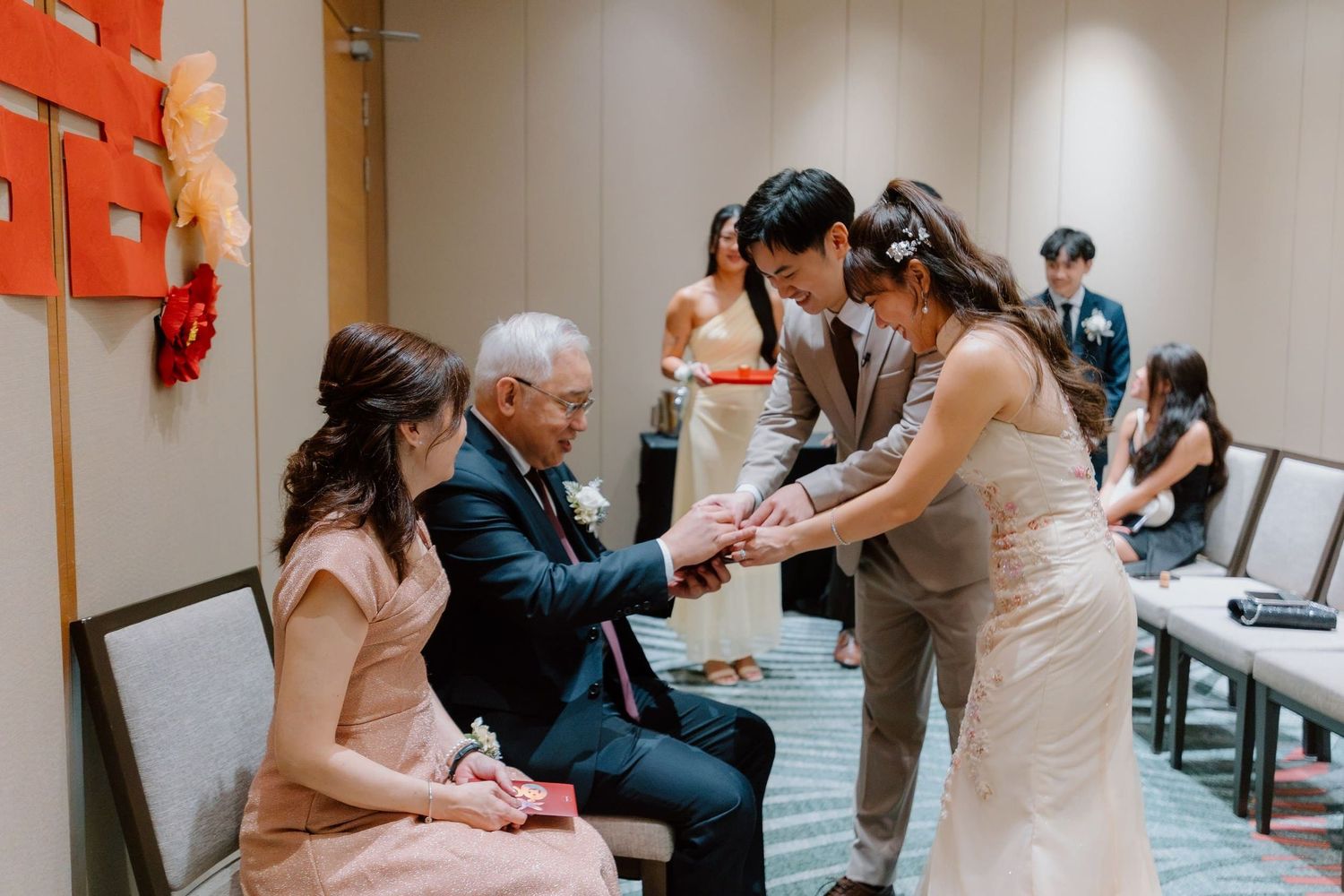 Elderly man seated with family members exchanging gifts during a traditional Asian wedding ceremony indoors.