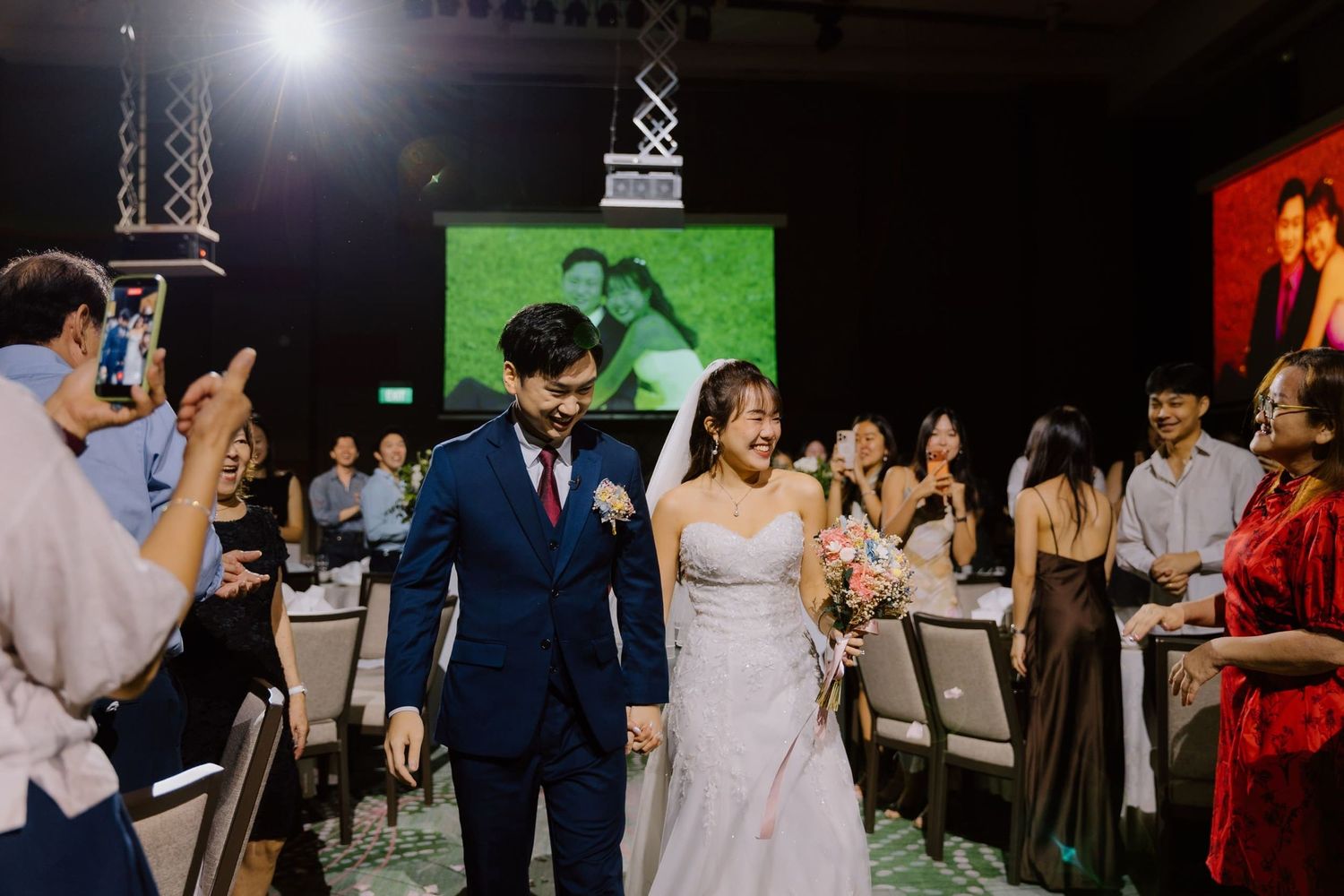 Bride and groom walk joyfully through guests at elegant wedding reception with green projection screen backdrop.