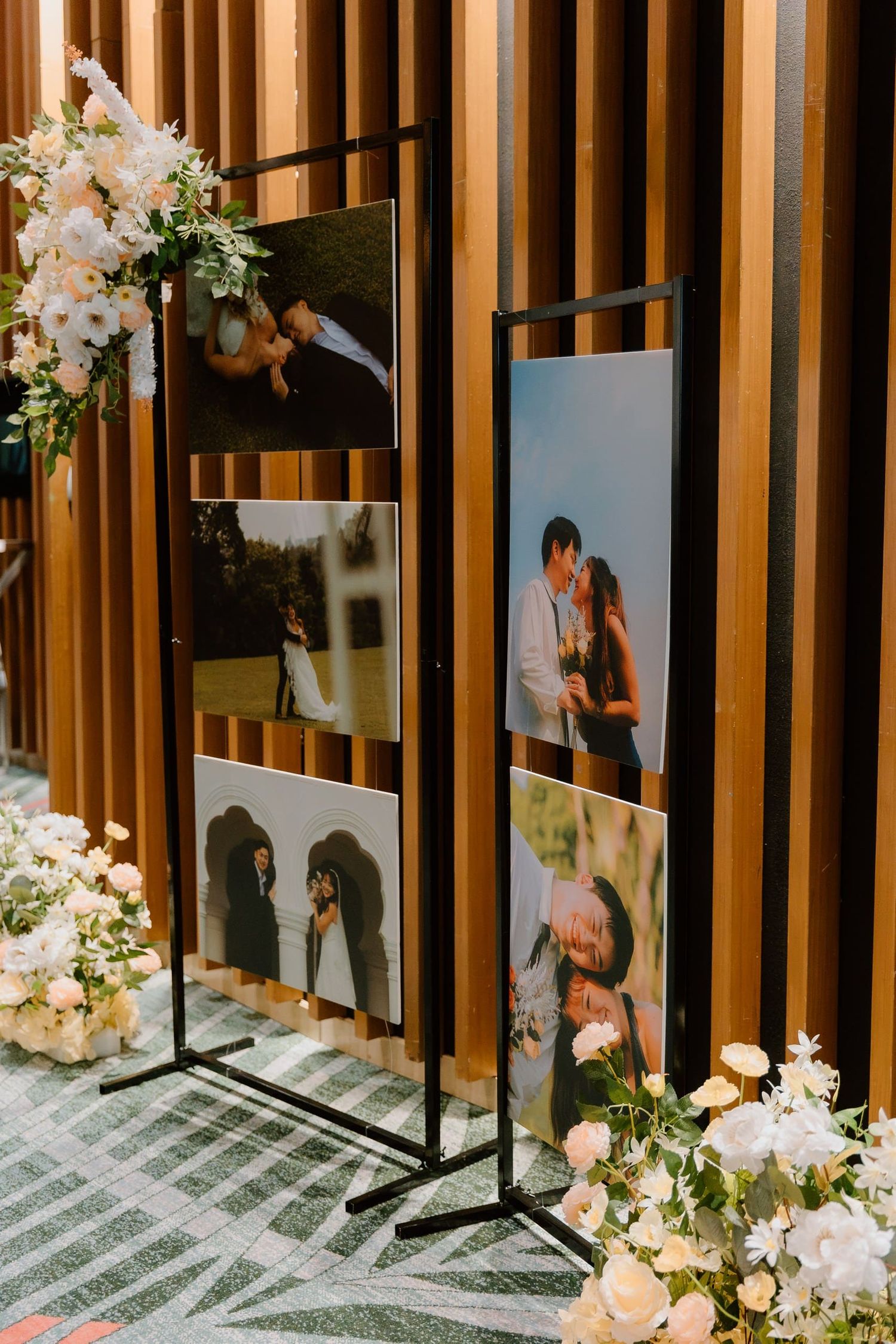 Black metal stands display wedding couple photos beside white floral arrangements against a wooden slat wall.