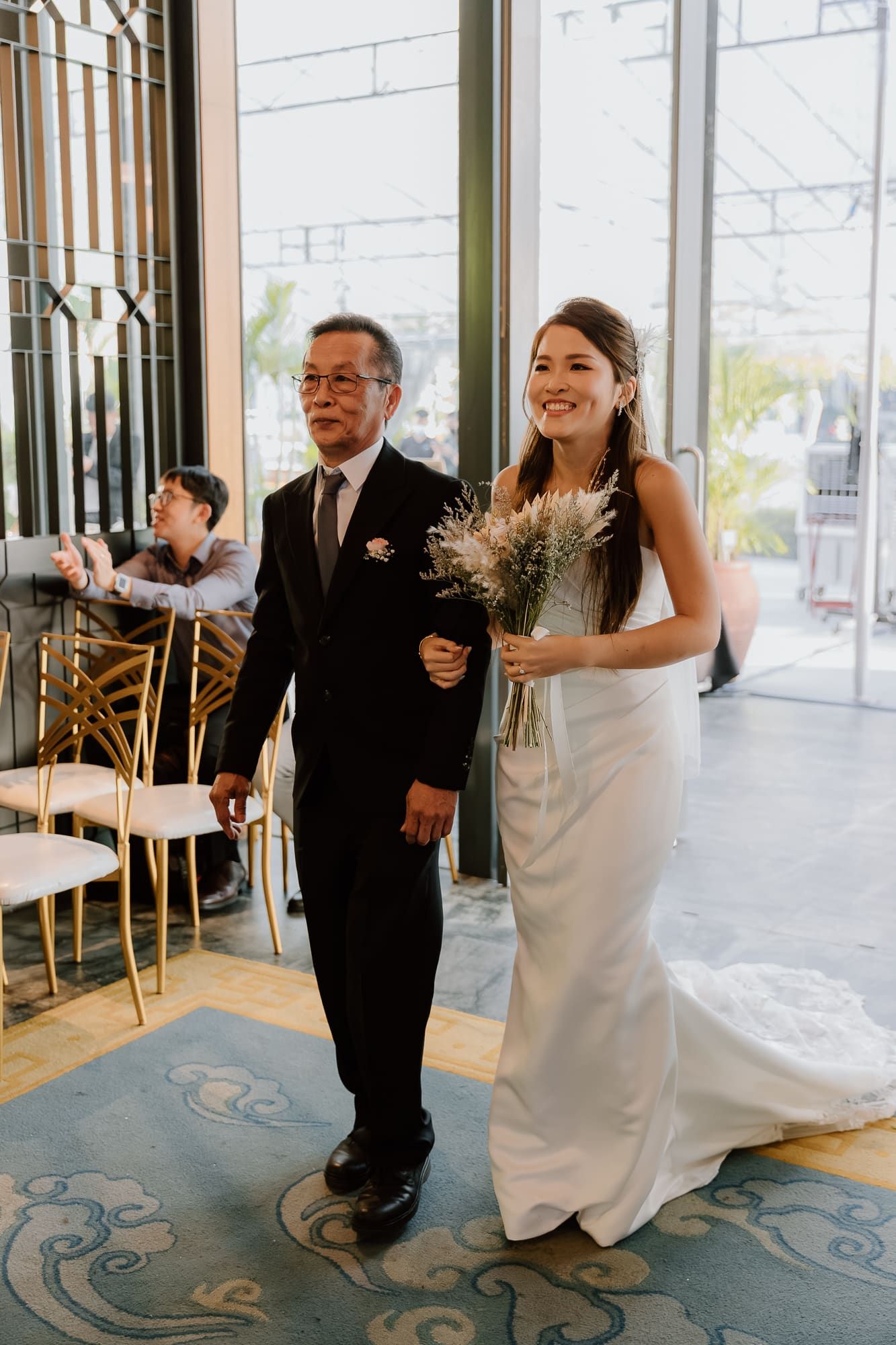 A wedding processional takes place in an elegant venue with ornate carpeting as the pair walks arm-in-arm down the aisle.