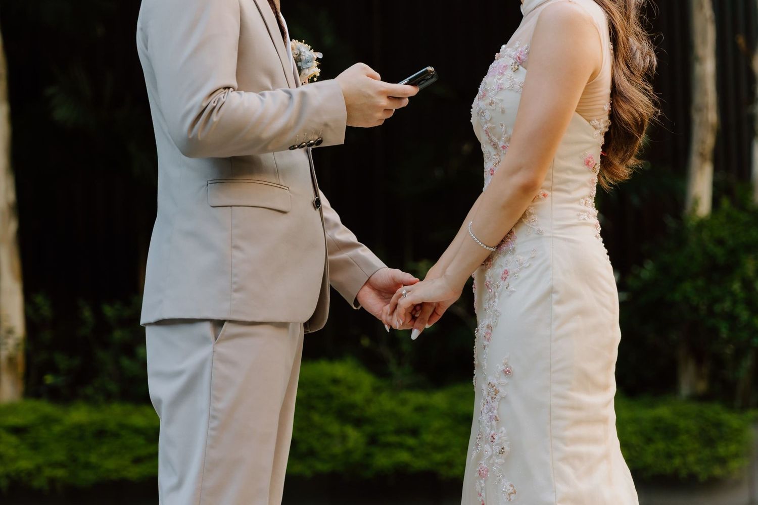 Bride and groom hold hands during wedding ceremony, groom in beige suit reads vows from phone.