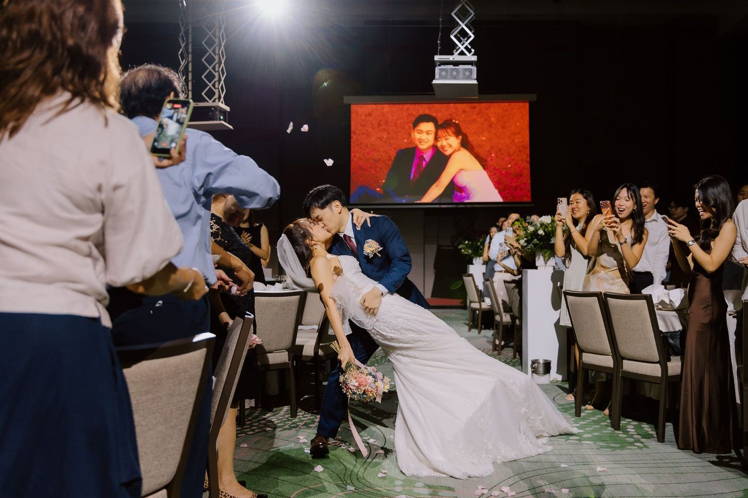 Bride and groom share a romantic dip kiss at wedding reception with guests applauding and couple photo displayed on screen.