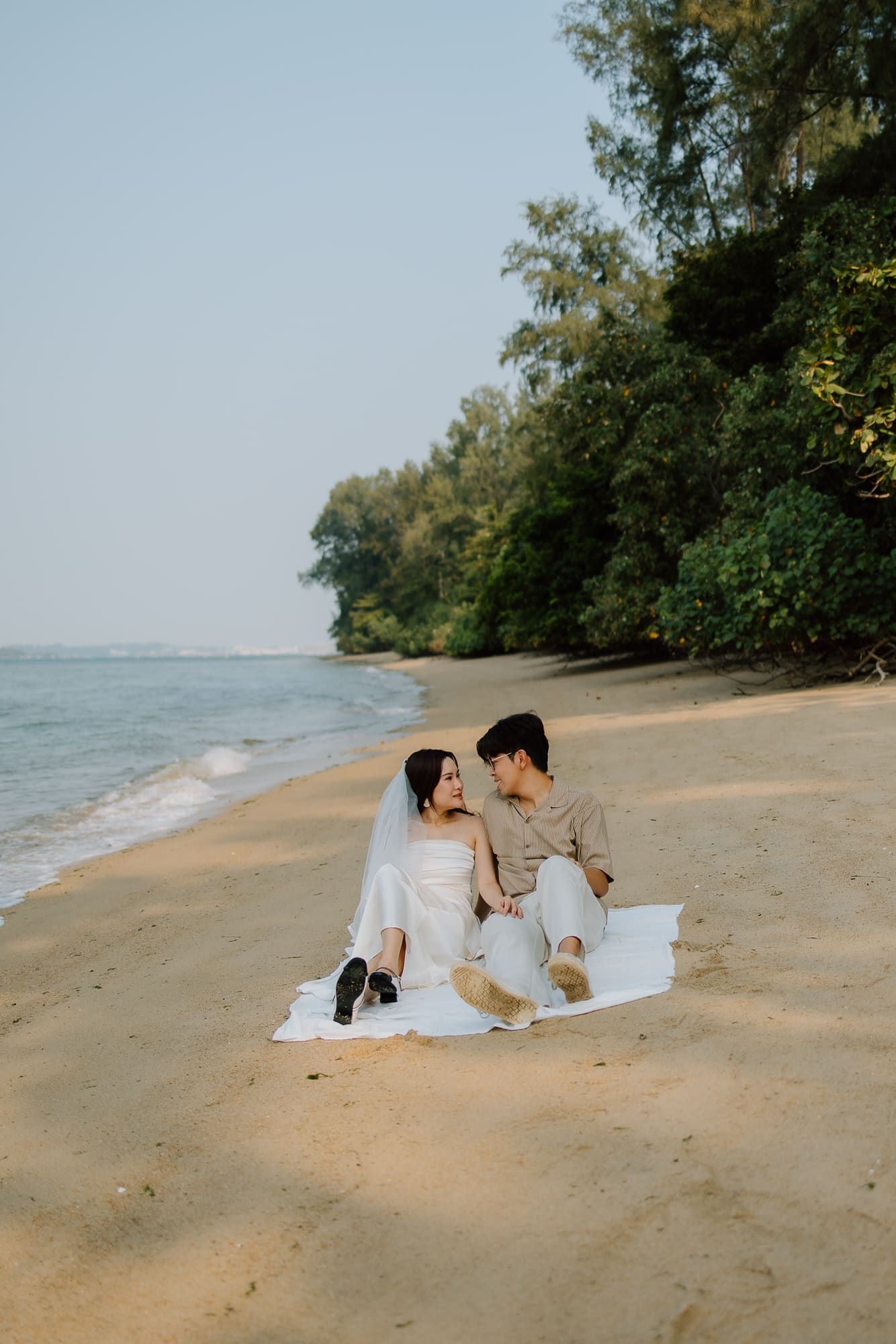 A couple in wedding attire sits romantically on a blanket along a tropical beach shoreline surrounded by lush green trees.