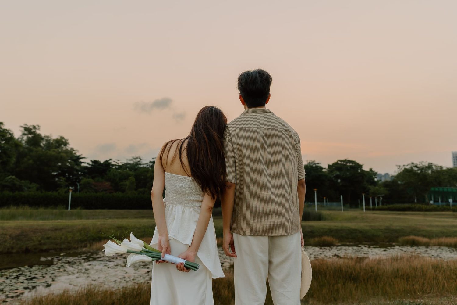 Couple holding flowers watching a pastel sunset over a green field, viewed from behind.