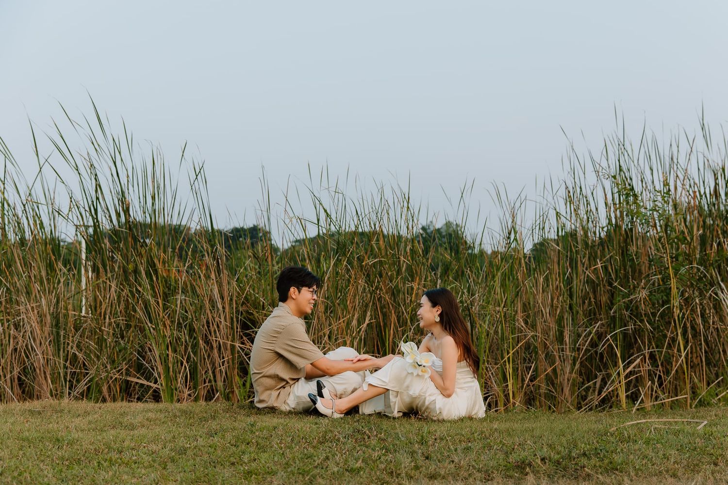 Couple sitting on grass in front of tall reeds, sharing a romantic moment outdoors in a serene natural setting.