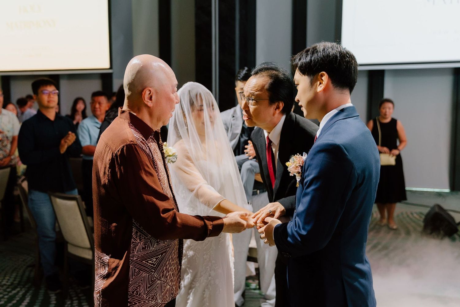 Asian wedding ceremony with bride in white veil, groom in suit, and officiant exchanging vows indoors.