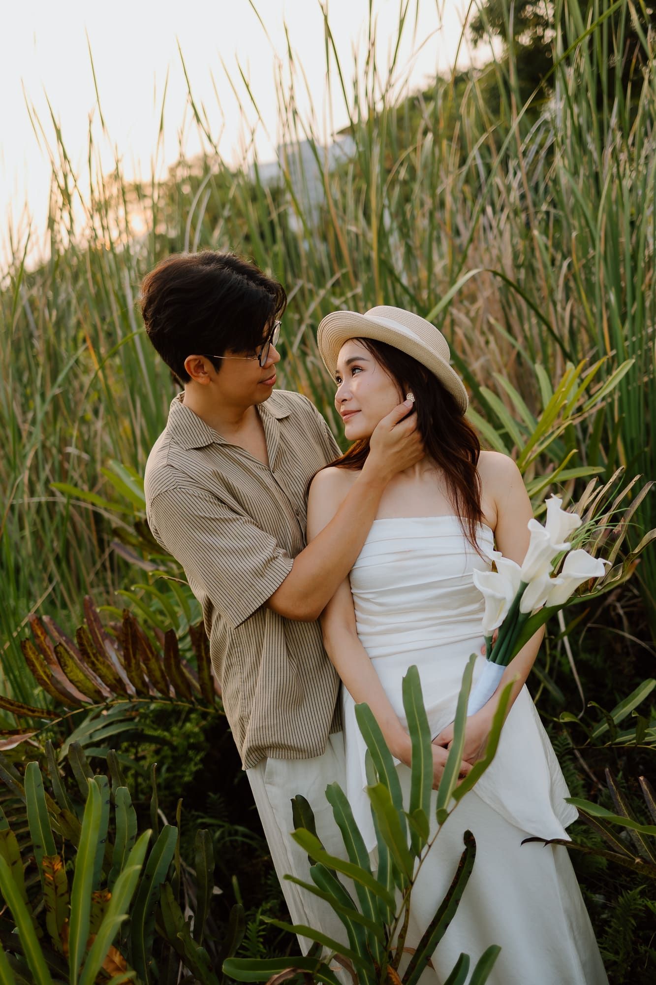 A couple shares a romantic moment in a lush green field, the woman in a white dress and hat holding flowers.