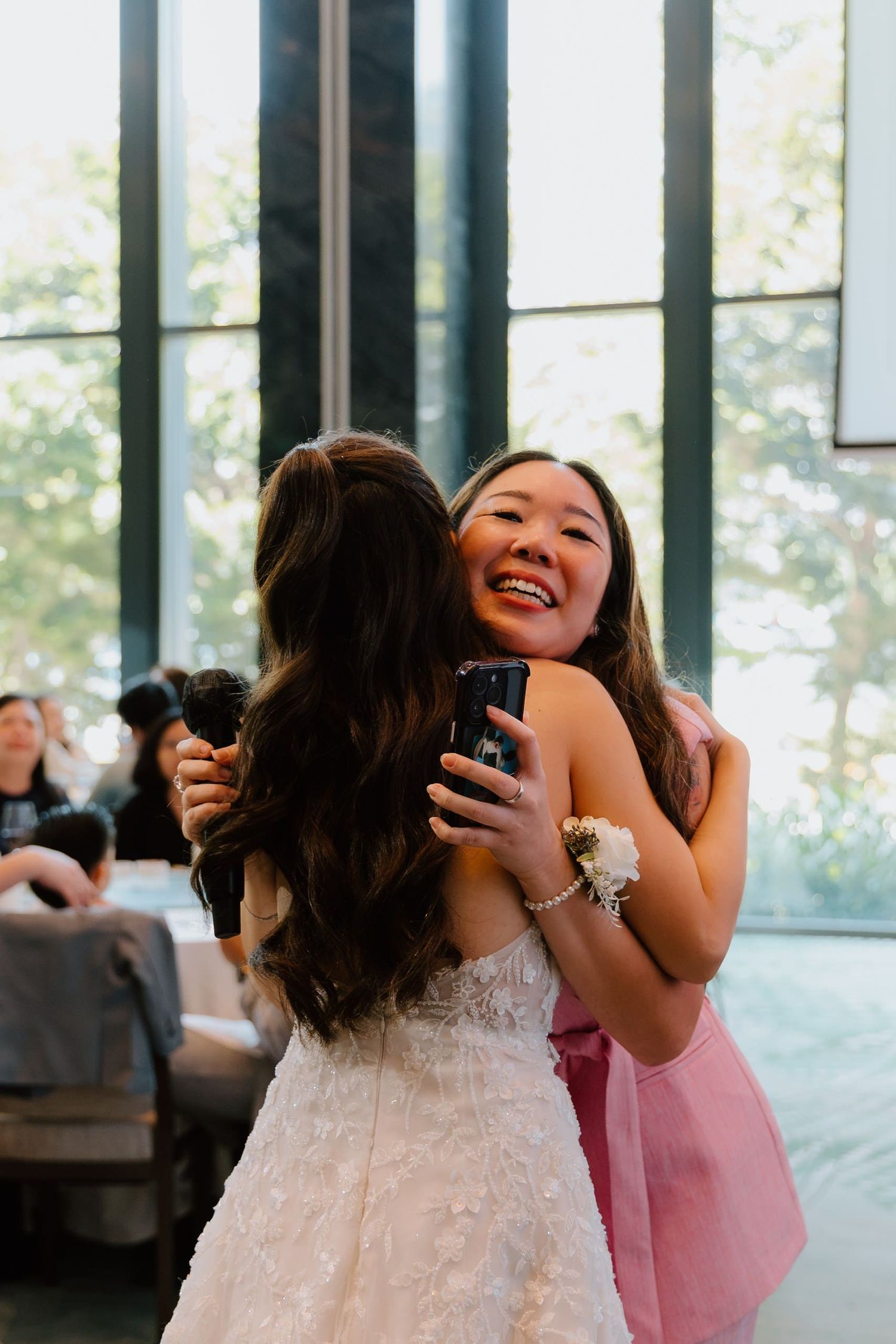 Two women share a joyful hug at a wedding reception, one in a white gown, the other in pink.