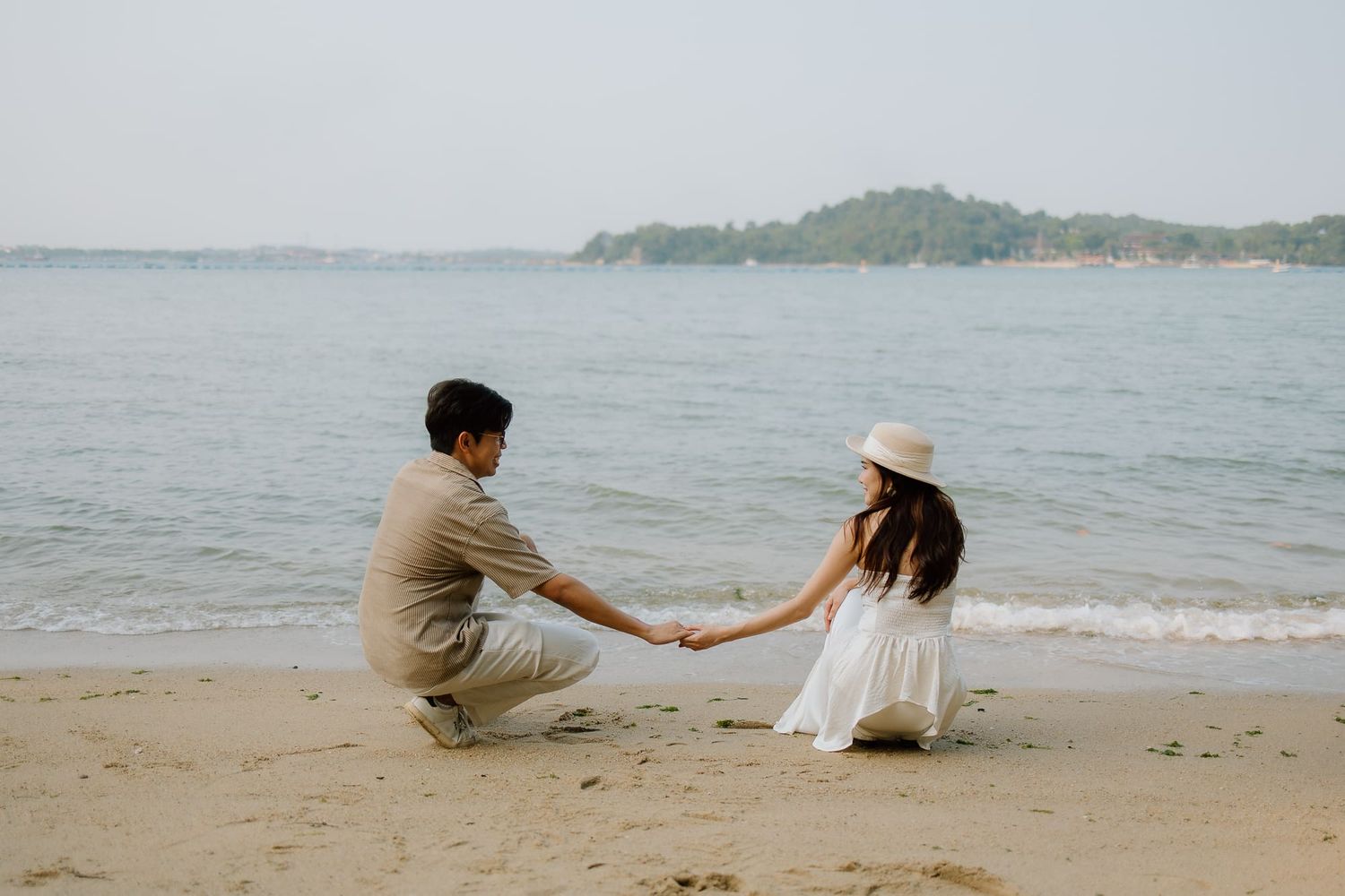 Couple holding hands on a sandy beach by calm blue water, woman in white dress and hat, romantic seaside moment.