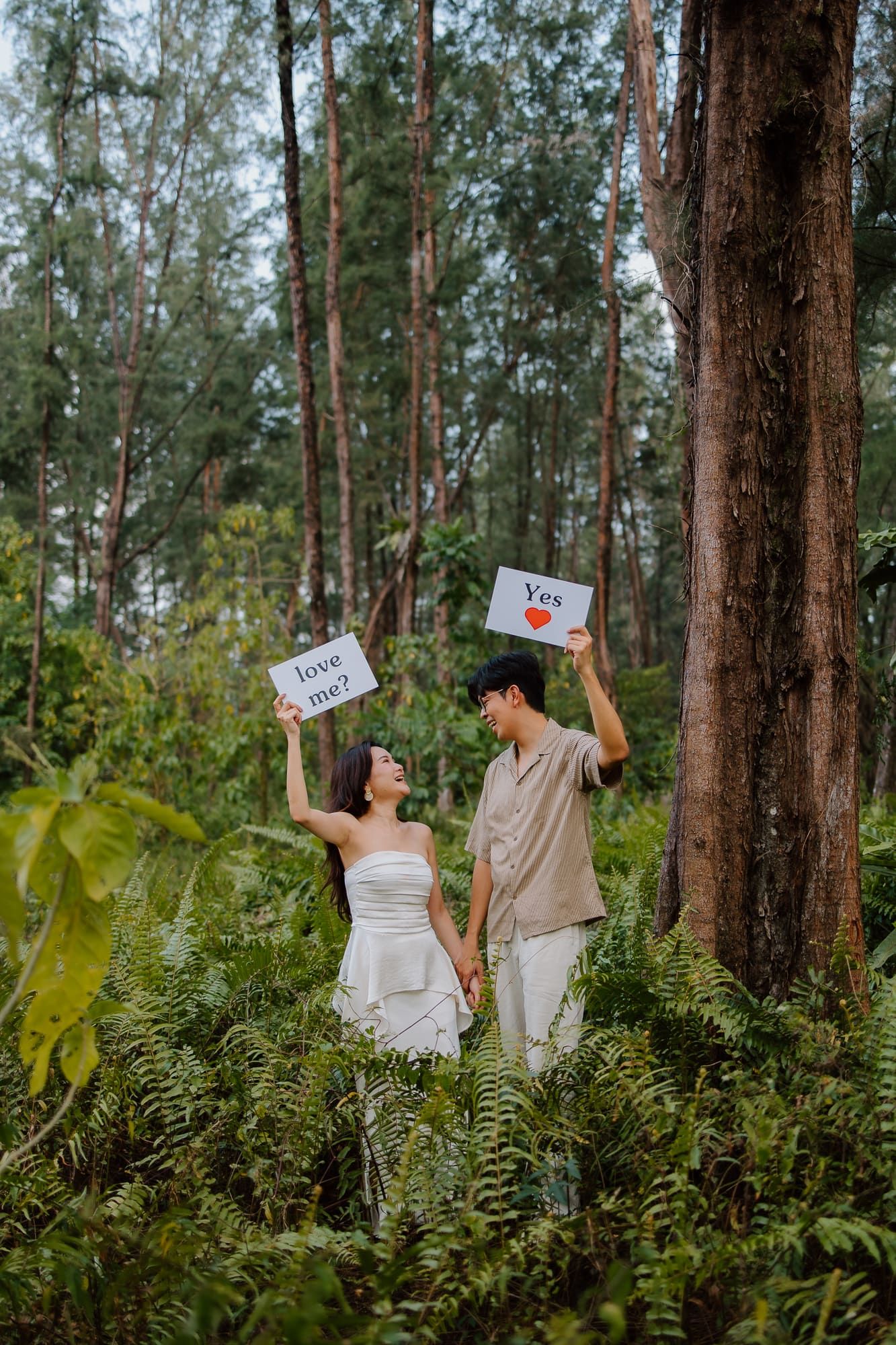 Couple holding 'love me?' and 'Yes' signs in a lush forest setting during a romantic outdoor proposal photoshoot.