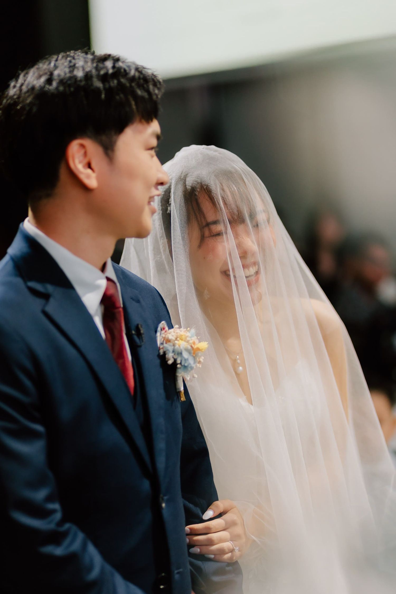 A smiling bride in a white veil and groom in a navy suit share a joyful moment during their wedding ceremony.