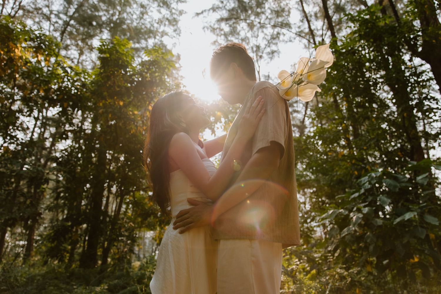 Couple embracing in sunlit forest with golden backlight filtering through lush green trees.