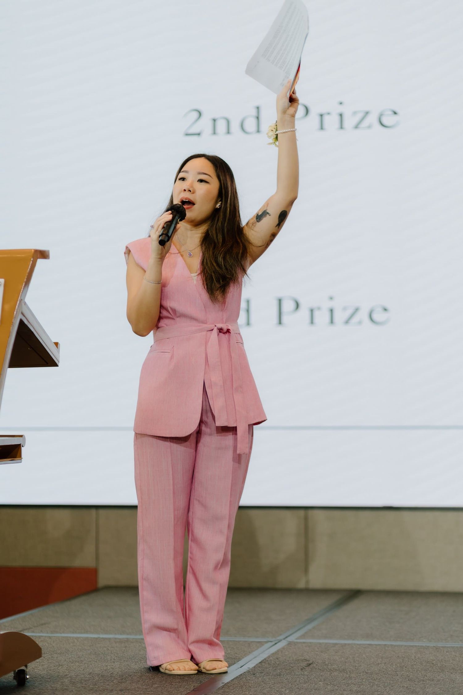 Woman in pink suit speaking on stage with microphone, raising paper hand, prize ceremony backdrop.