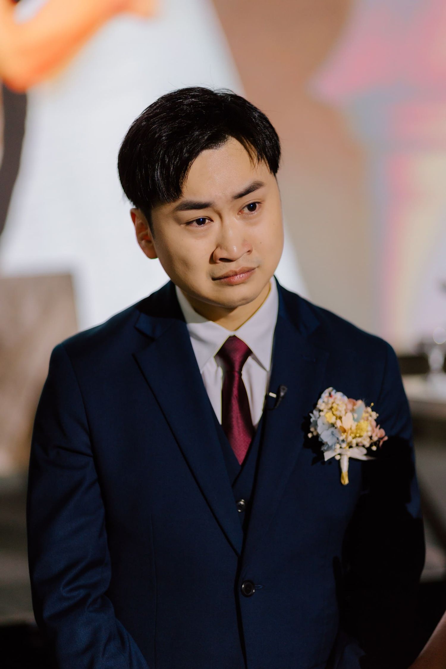 Emotional groom in navy suit with floral boutonniere and red tie at wedding ceremony.