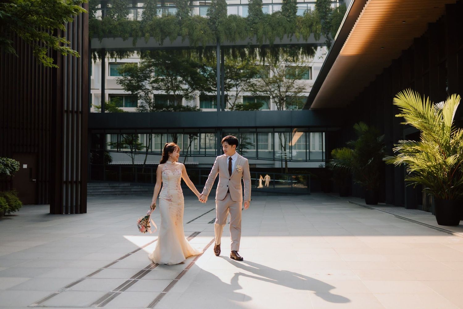 Bride and groom holding hands walking through modern outdoor venue with lush greenery and contemporary architecture.