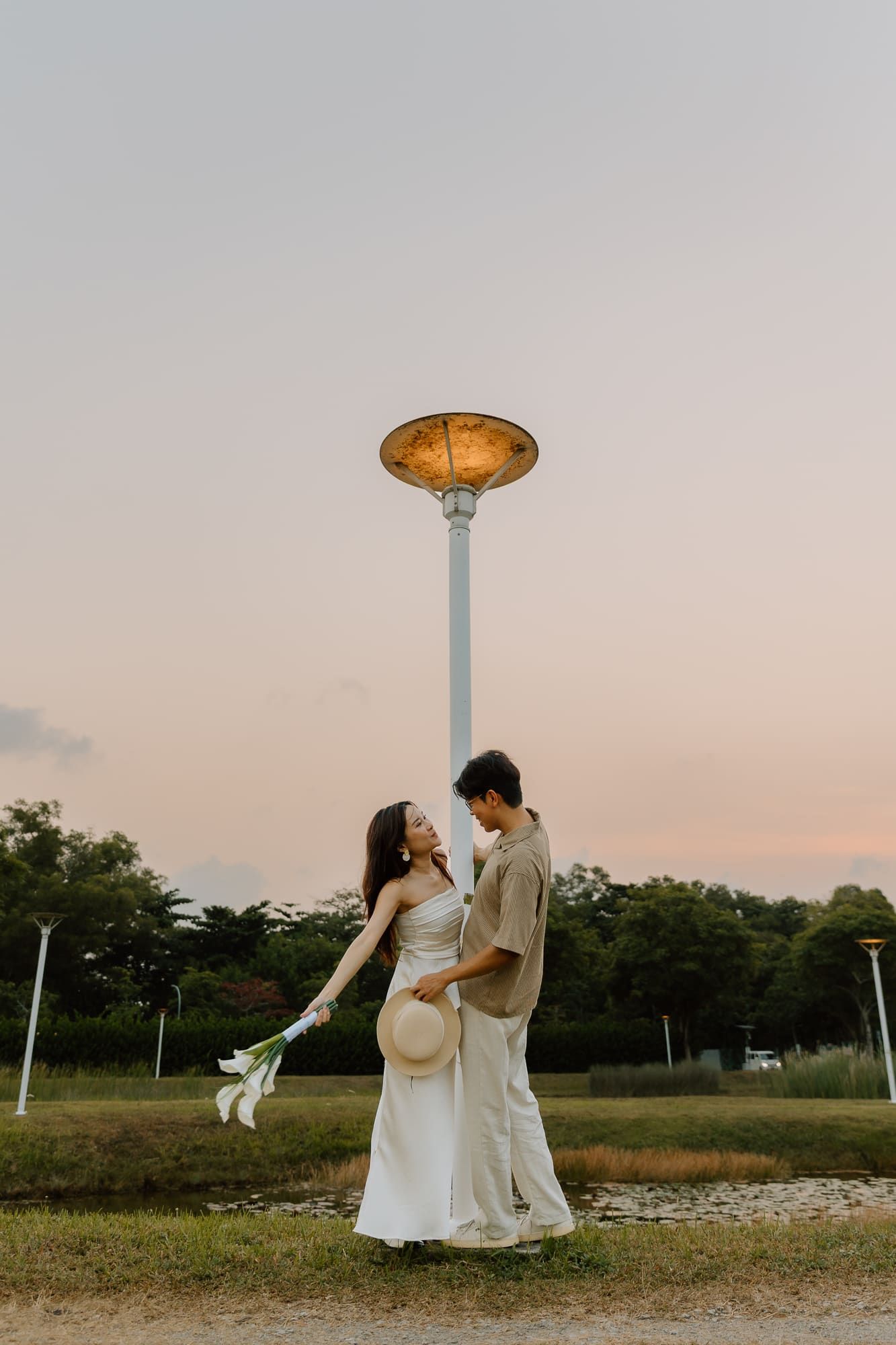 Couple in white and beige outfits holding flowers and a hat, embracing at sunset near a tall lamp post in a park.