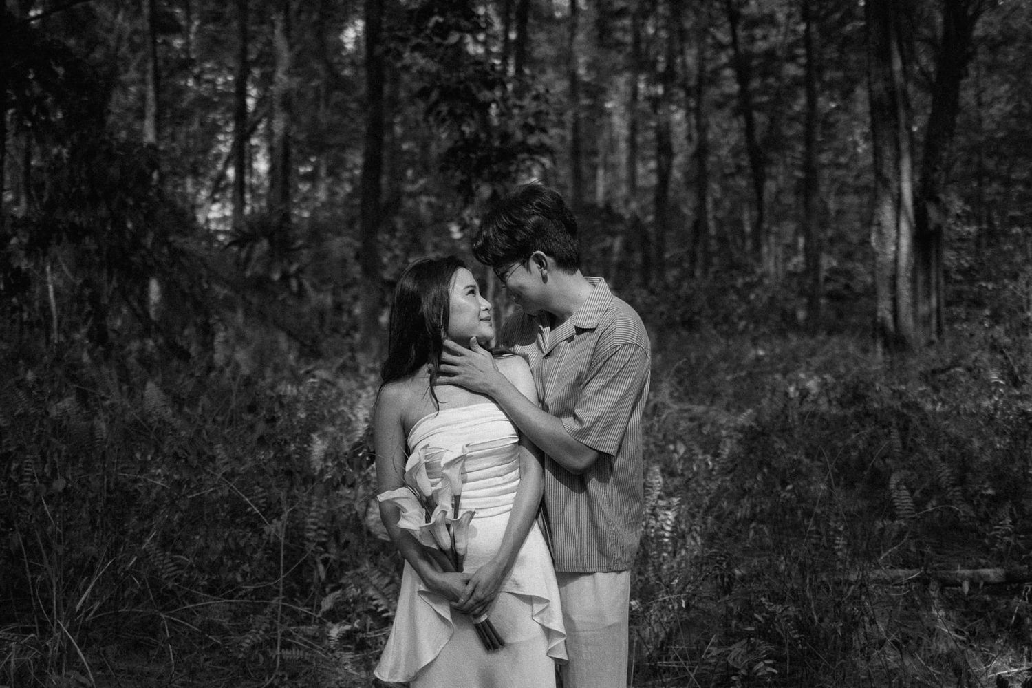 Black and white photo of a romantic couple embracing tenderly in a lush forest setting.