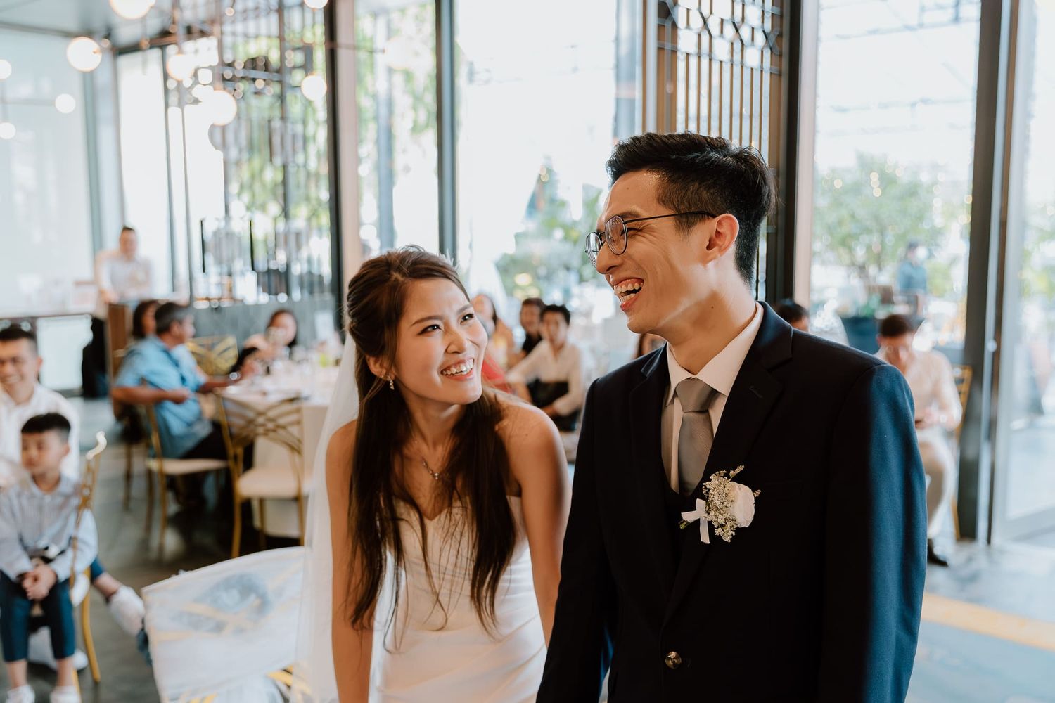 A happy couple shares a joyful moment together at their wedding reception in a bright, window-filled venue.