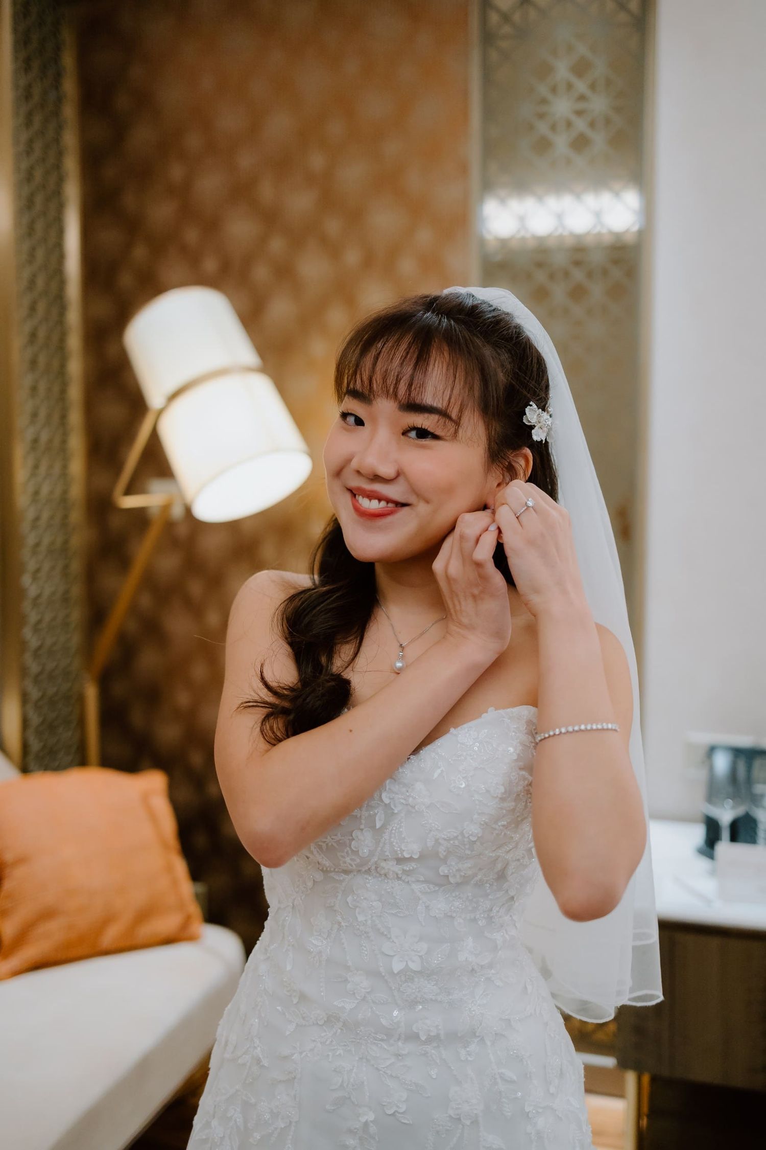 A smiling bride in a white strapless gown adjusts her earring while wearing a veil and pearl jewelry.