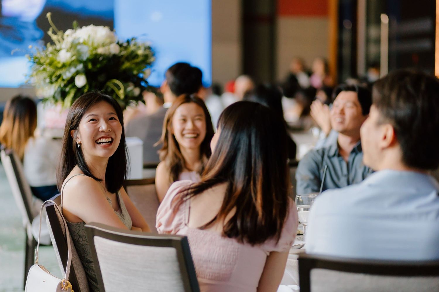 Guests smiling and laughing at a formal dining event with floral centerpieces in a bright modern venue.