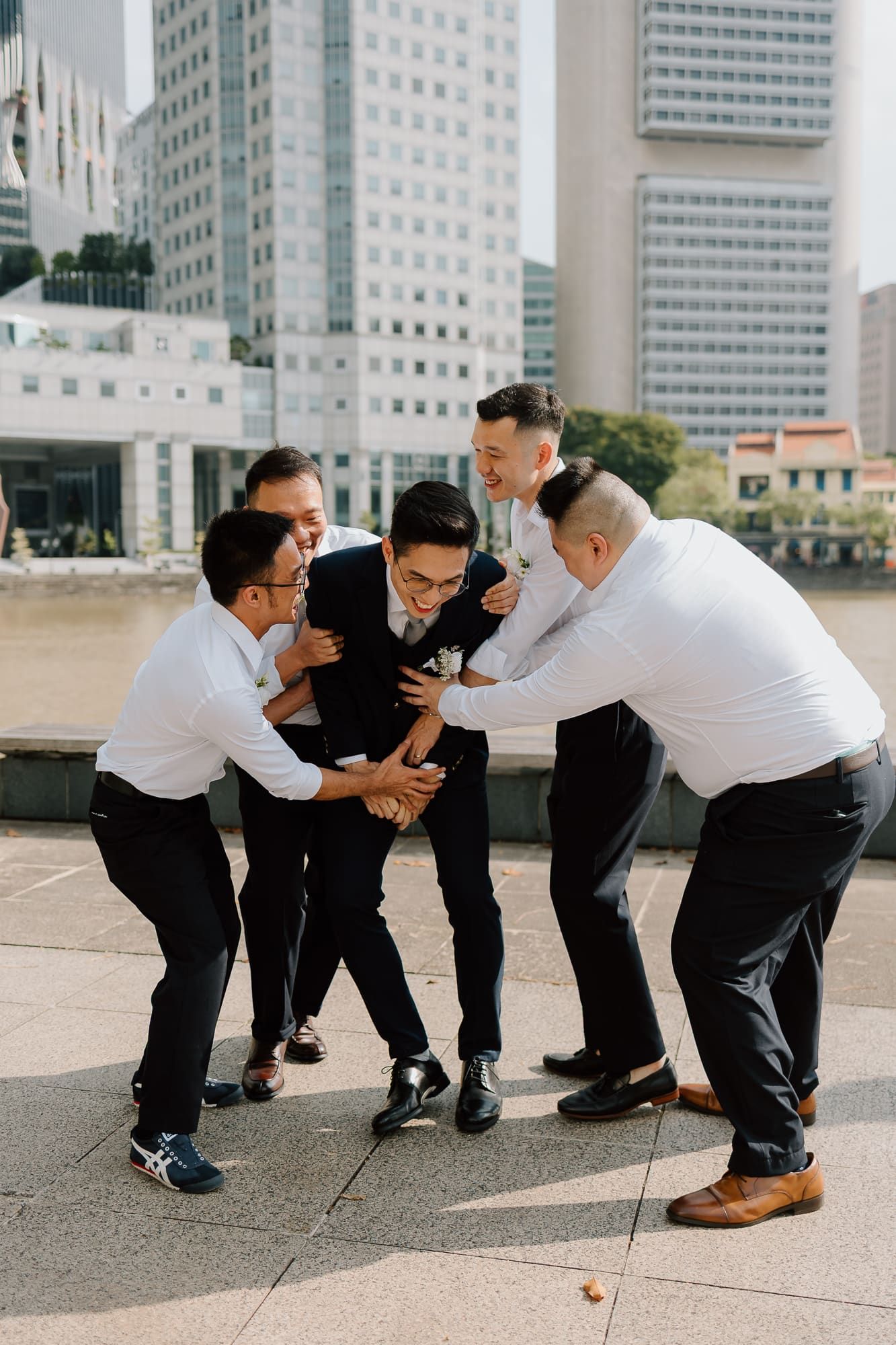 Group of friends in business attire playfully huddle together and laugh on city sidewalk with skyscrapers in background.