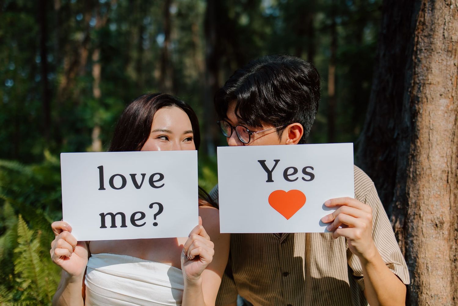 Couple holding signs reading love me and yes with heart outdoors in forest setting.