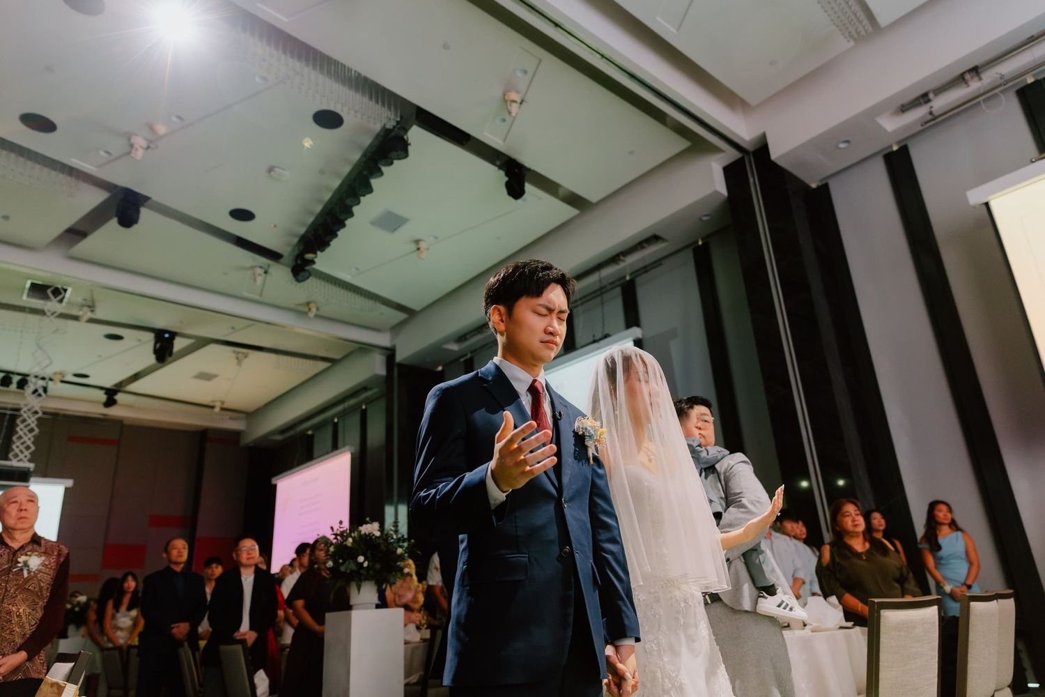 Groom in navy suit walks bride in white veil down the aisle at an elegant indoor wedding ceremony.