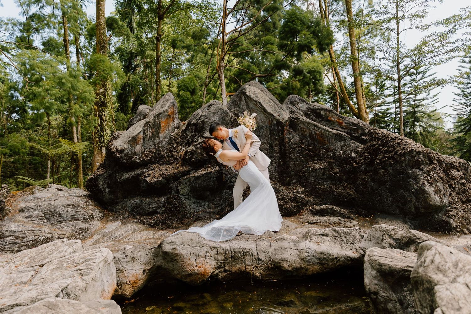 A couple in wedding attire poses for romantic photos among tall pine trees in a forest setting.