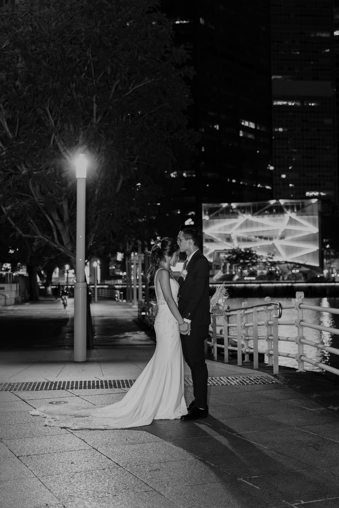 A romantic nighttime cityscape with a couple in formal wedding attire standing under a glowing street lamp.