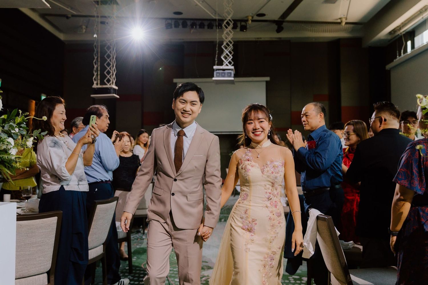 Happy Asian couple walks hand-in-hand at their wedding reception while guests applaud in elegant venue.