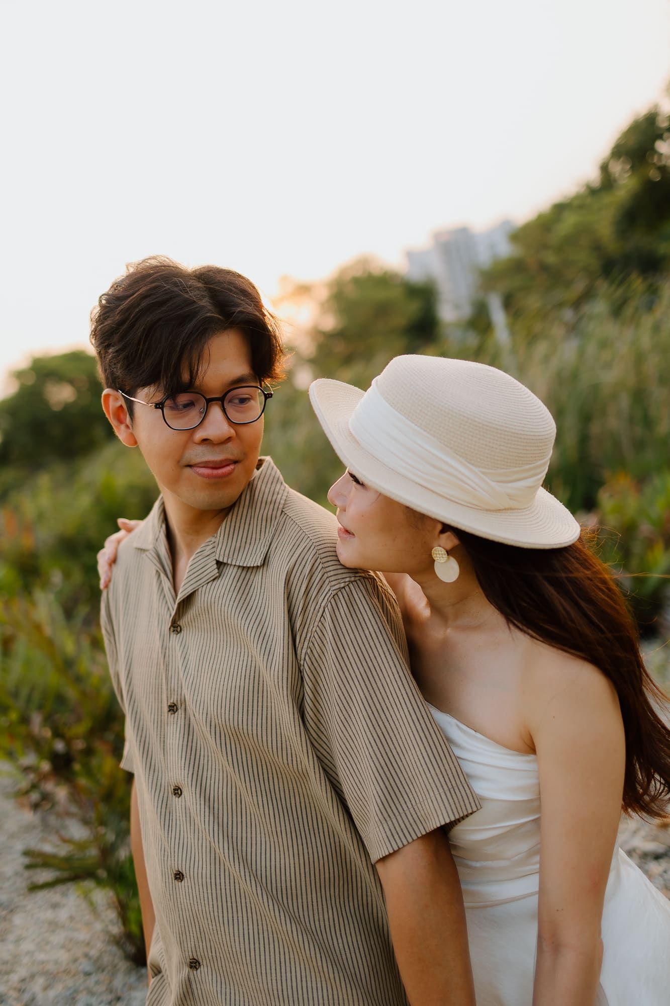 A couple poses outdoors at sunset, woman in white dress and hat leans on mans shoulder in a romantic portrait.
