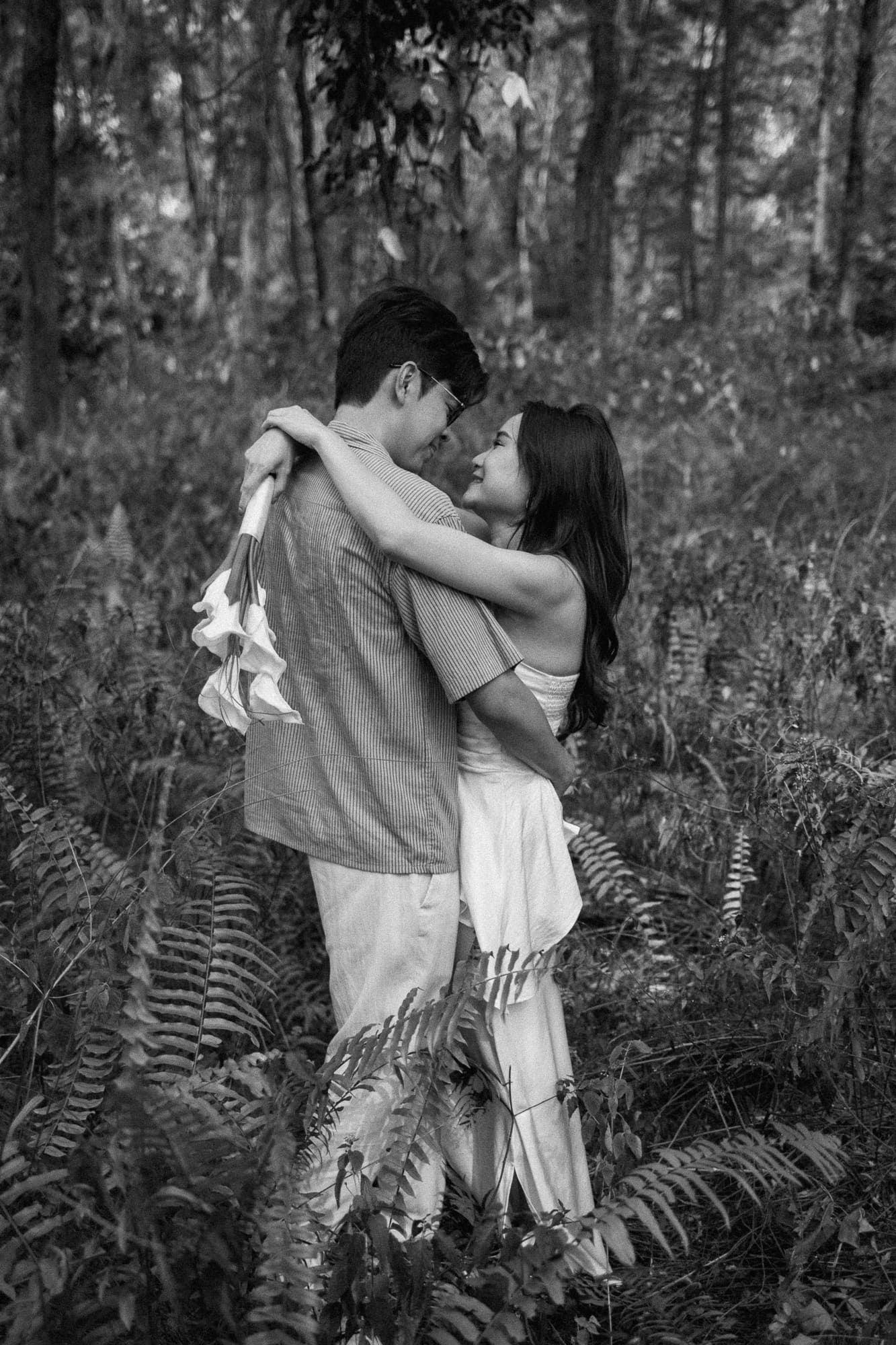 Black and white photo of a couple embracing romantically in a lush forest surrounded by ferns.