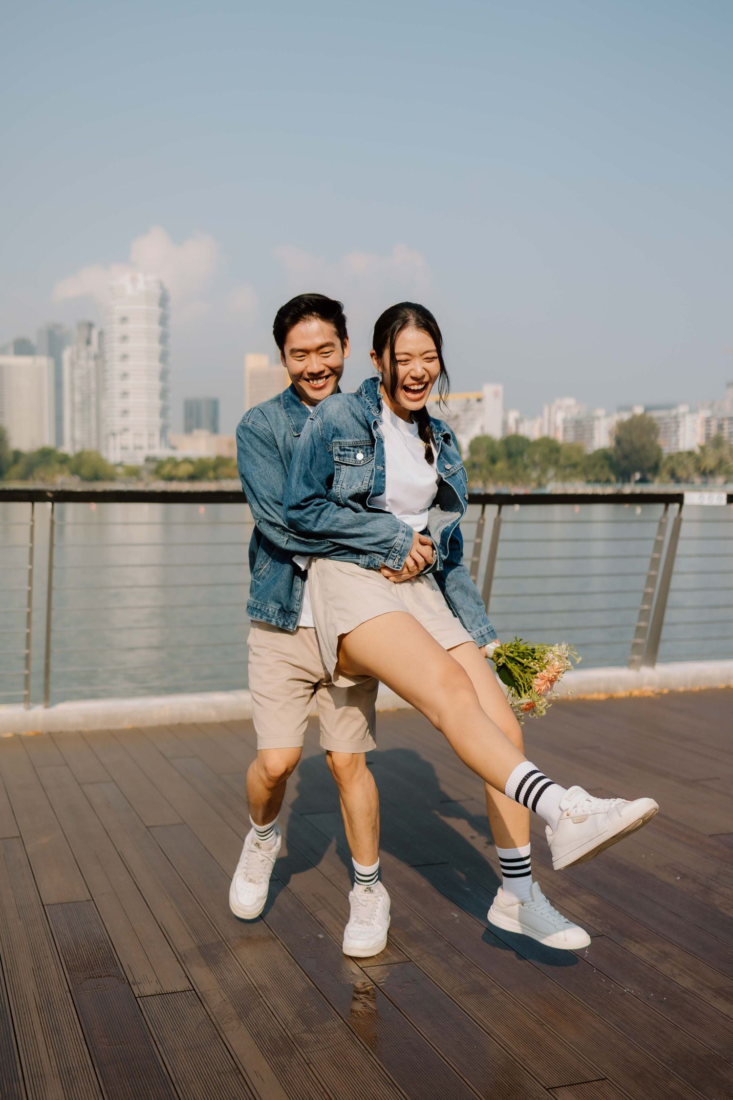 Happy Asian couple in matching denim jackets laughing on waterfront boardwalk, woman holding flowers, city skyline background.