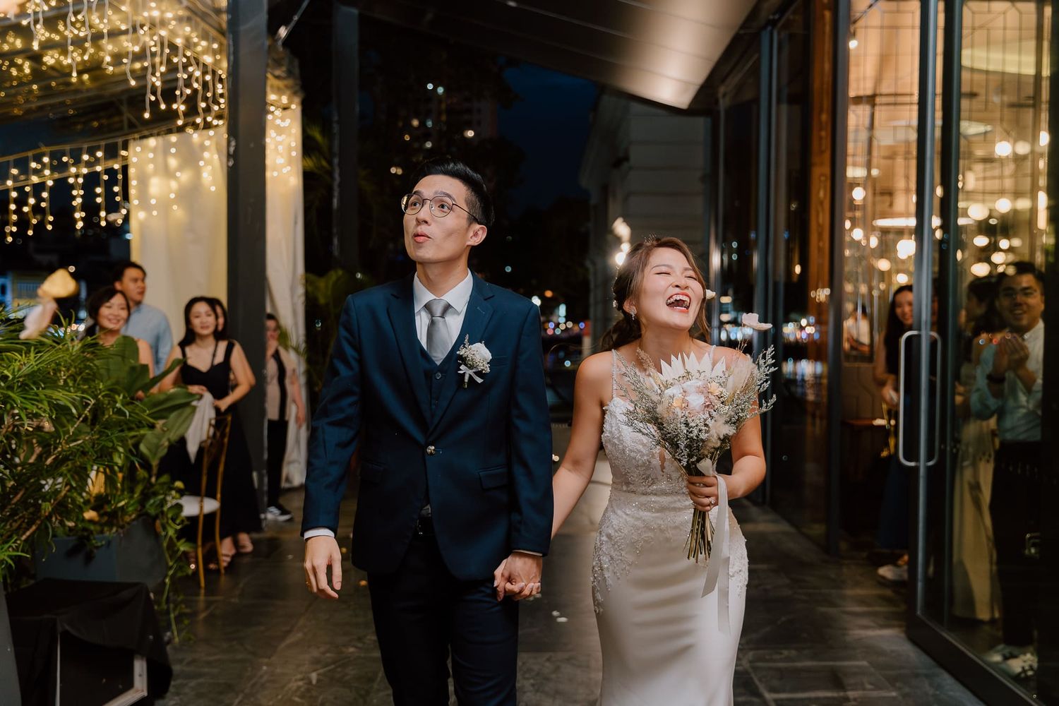 Newlywed couple laughing while exiting venue with decorative lights and greenery at evening reception.