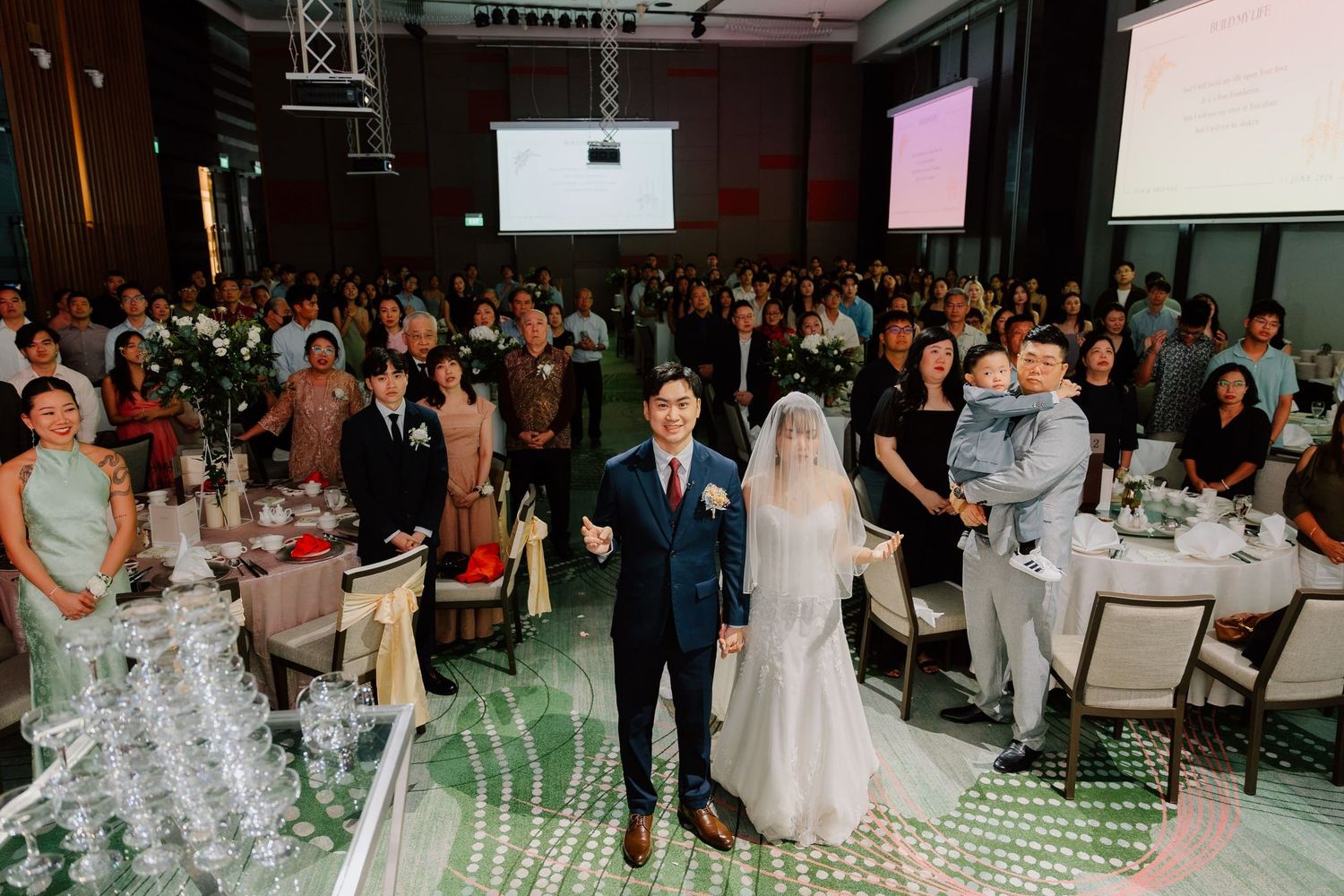 Bride and groom stand at the aisle of an elegant wedding reception hall filled with guests at round tables.