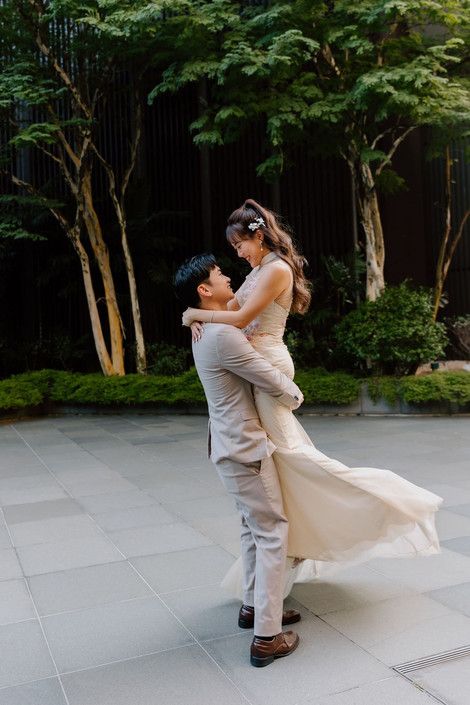 A groom lifts his bride in a flowing champagne gown outdoors, surrounded by lush green trees on a tiled plaza.
