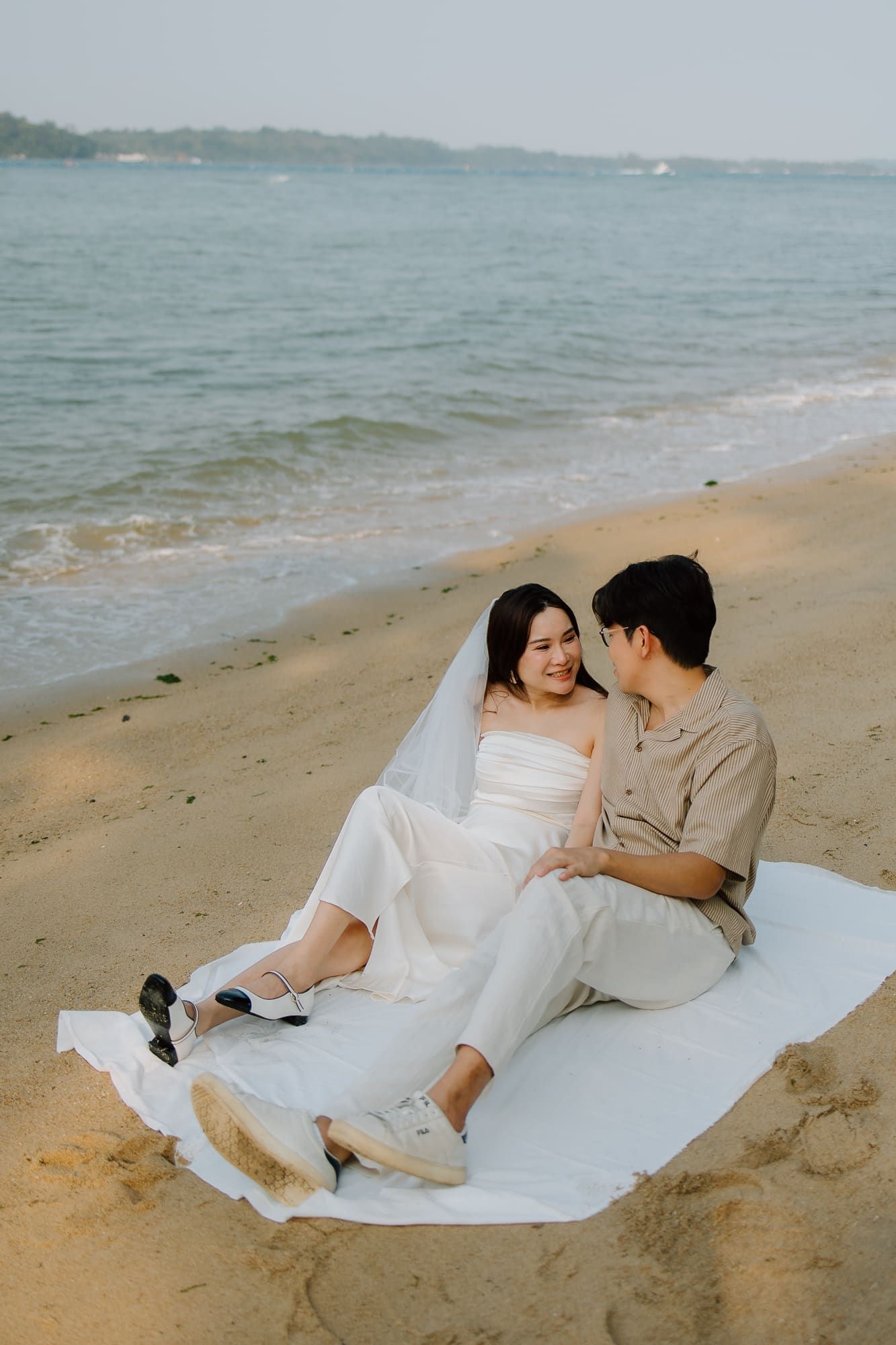 A couple in wedding attire sits on a white blanket on a sandy beach, gazing at each other near the ocean.