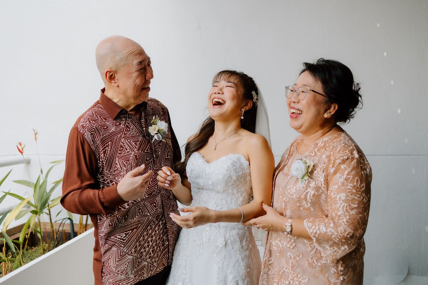 Bride in white gown laughing joyfully with parents wearing floral outfits at a wedding celebration.