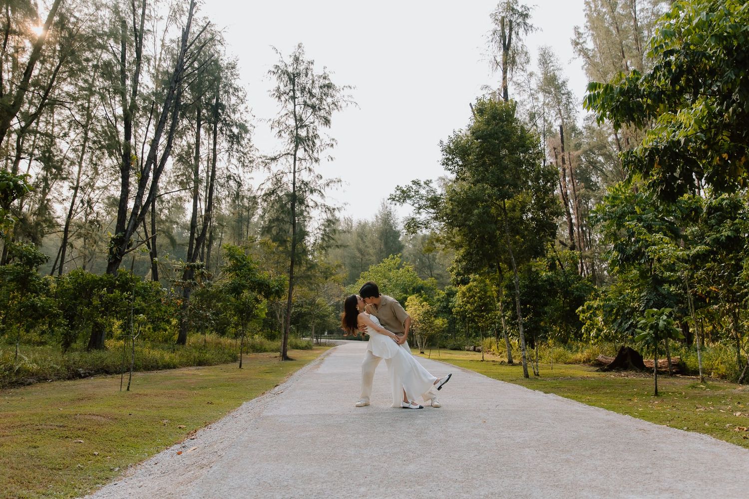 Couple sharing a romantic dip kiss on a tree-lined path in a lush green park during their wedding day.