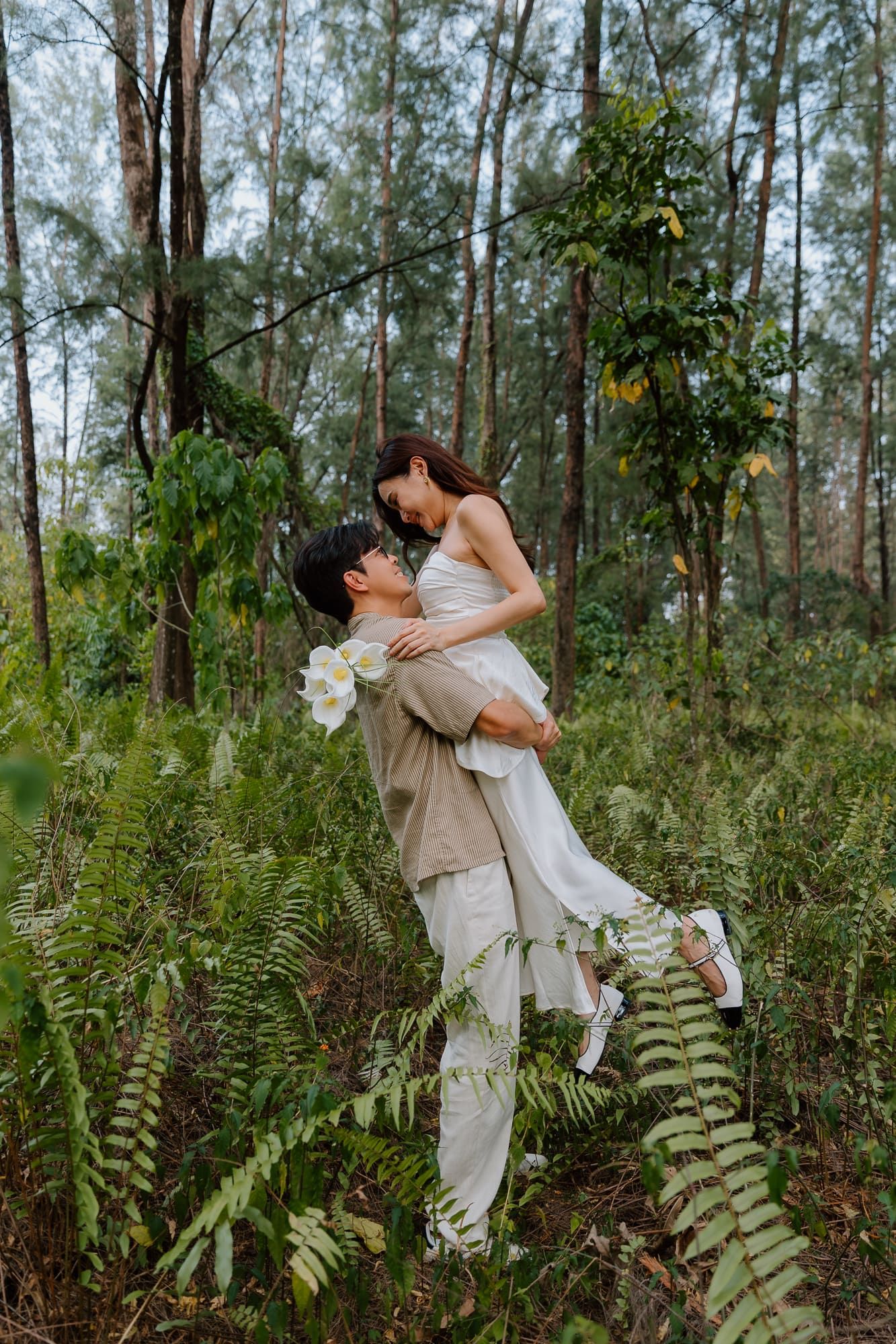 A groom lifts his bride in a lush forest setting, both dressed in elegant white and beige wedding attire.