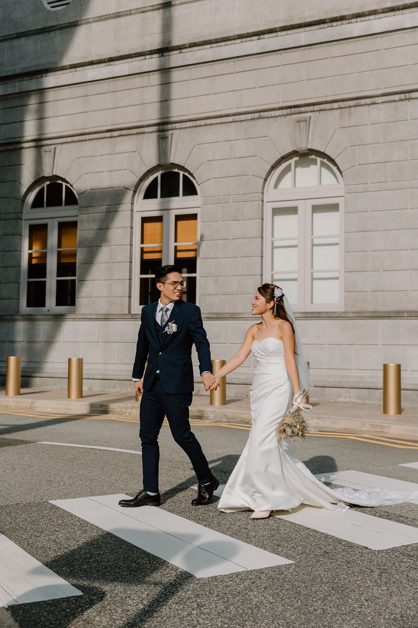 Wedding couple playfully poses on concrete pavement with white building facade in background.