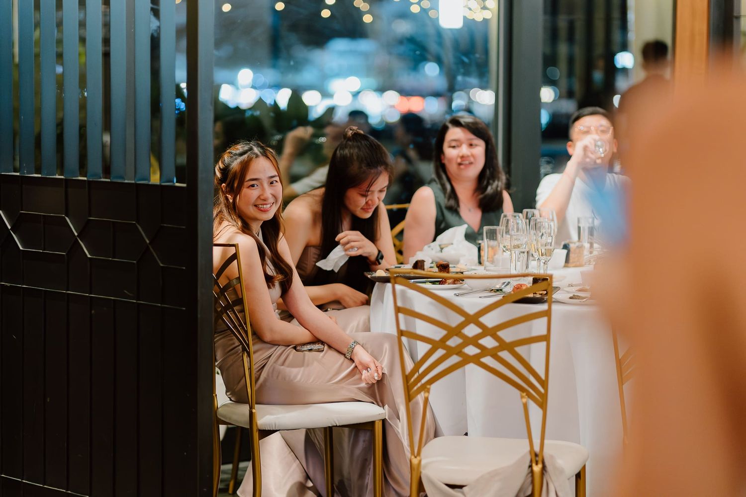 Wedding party members in light colored attire gather around an elegantly decorated glass table at a reception.