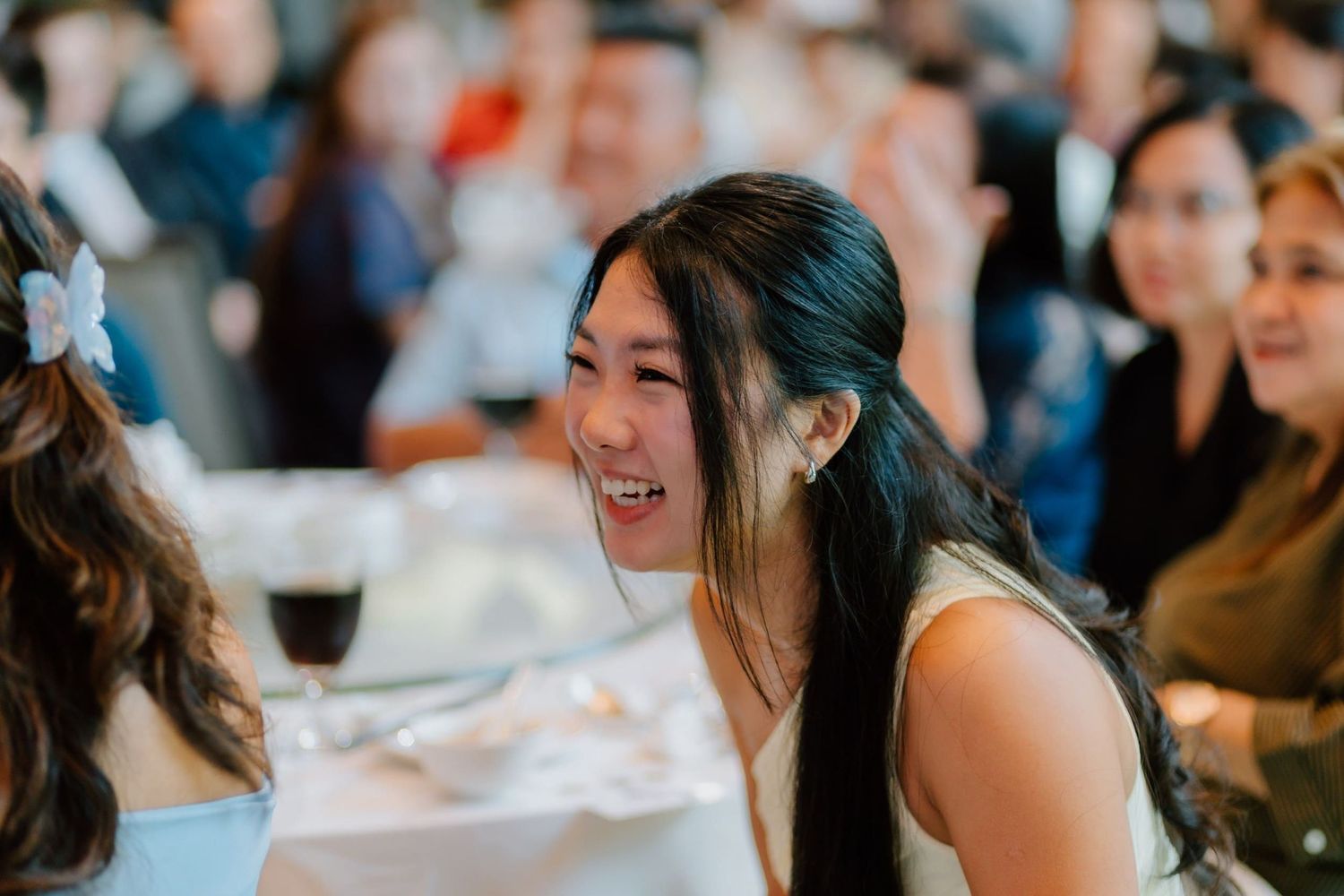 A joyful young woman with long dark hair laughs at a wedding reception dinner table.