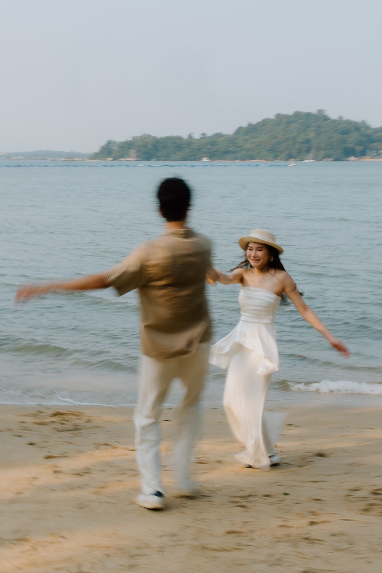 A couple holds hands and dances joyfully on a sandy beach, with the woman wearing a white dress and sun hat.