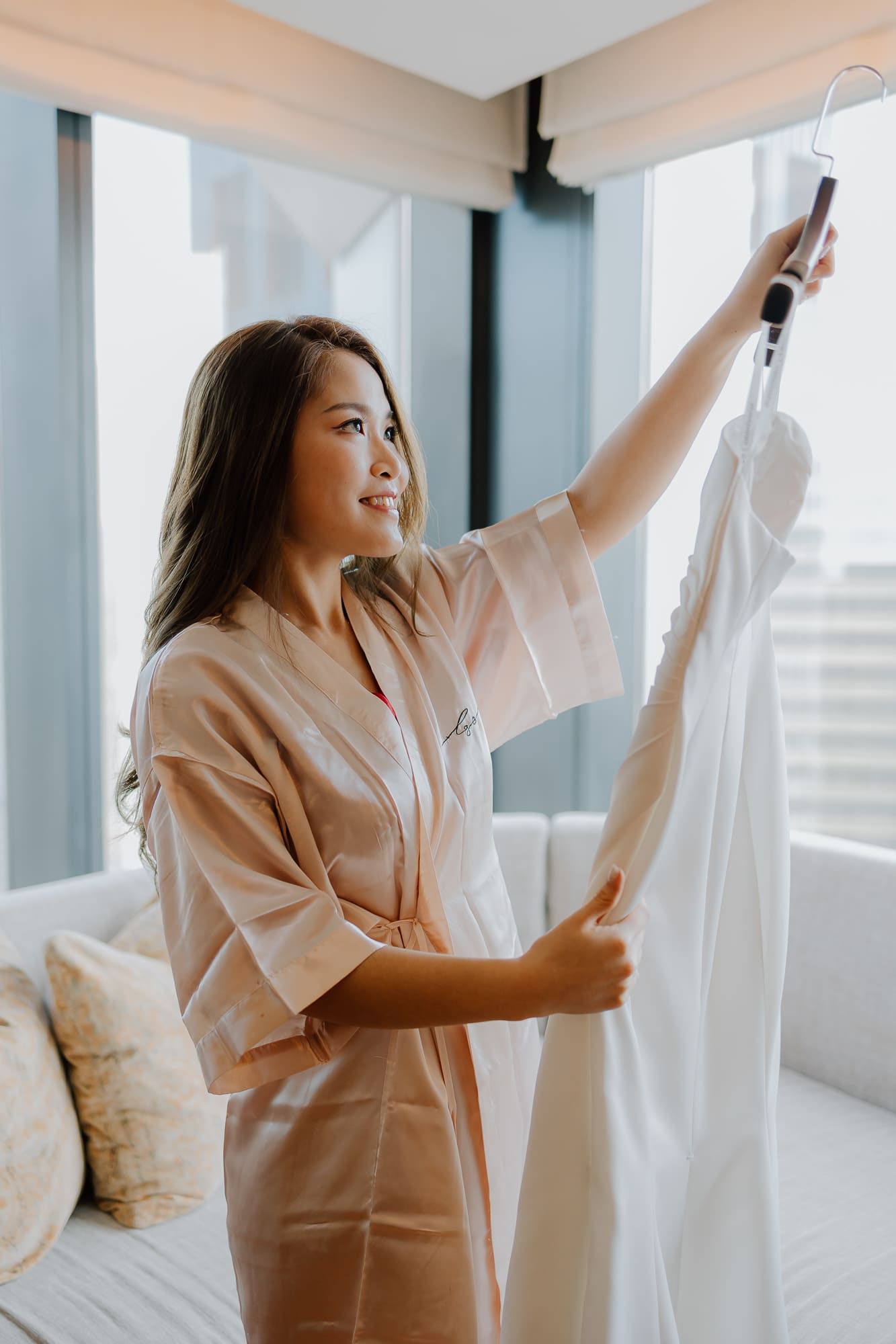 Woman in beige robe getting ready in bright modern room with windows while hanging up white clothes.